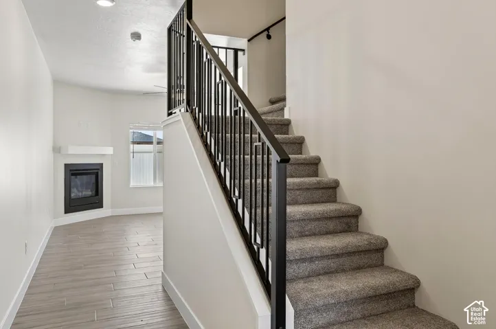 Open-concept living area featuring a fireplace with a white mantle, wood-finish flooring, and a carpeted staircase with a black metal railing
