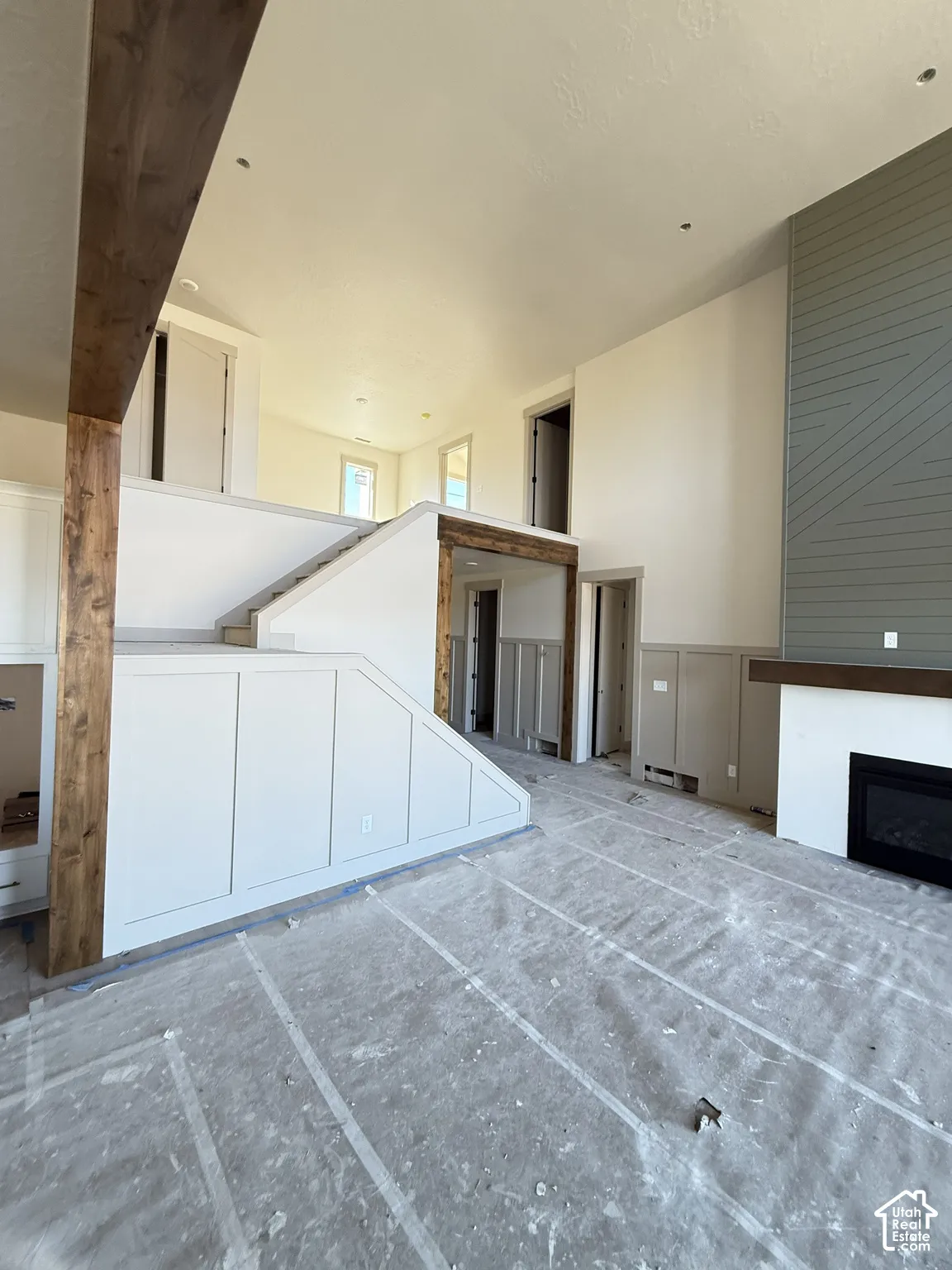High-ceiling living area featuring a floor-to-ceiling fireplace with horizontal wood-panel detailing, exposed structural wood beams, custom shaker-style wall paneling, and a staircase with integrated under-stair cabinetry