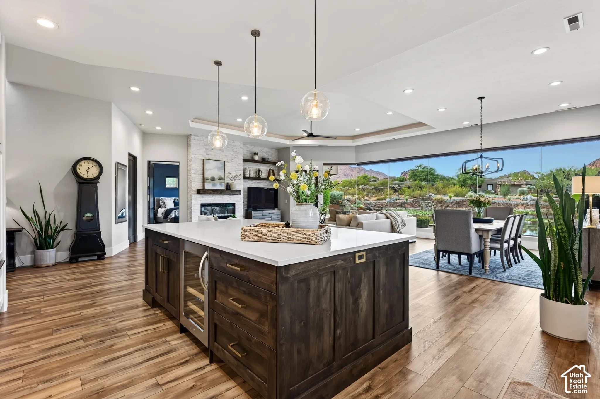 Open-concept living area with wood-finish flooring, a kitchen island with built-in wine refrigeration, and a fireplace with a stacked stone surround