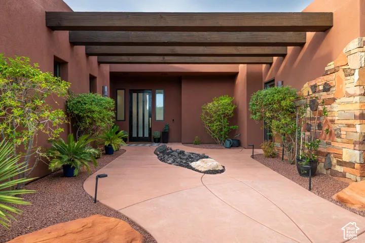 Entryway featuring an architectural wood beam trellis, a modern glass-paneled entry door, a curved concrete pathway, and decorative stone landscaping