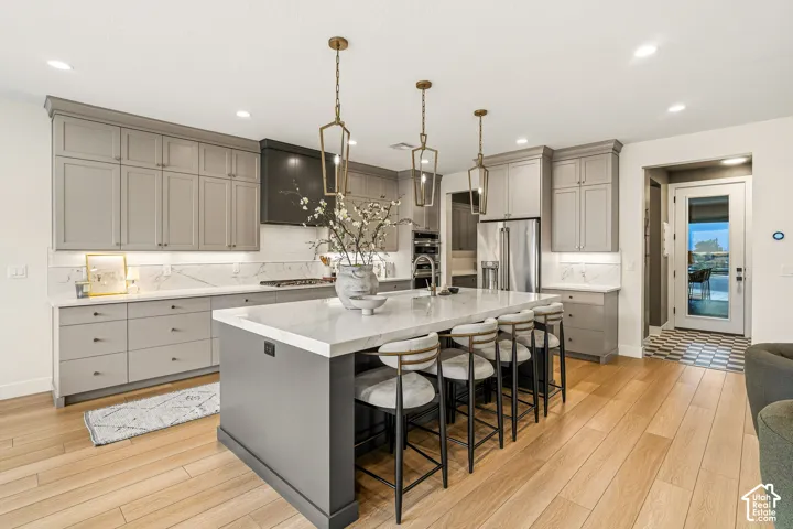 Expansive kitchen featuring light wood-finish flooring, a large island with white stone countertop, grey cabinetry, stainless steel appliances, and decorative pendant lighting