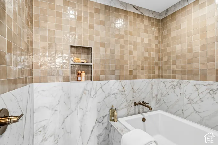 Built-in soaking tub with a wall-mounted bronze faucet, marble-look paneling, and tile surround with a recessed shelf