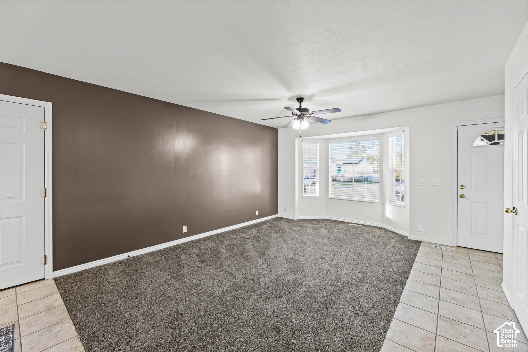Spacious room featuring gray carpeting, a bay window with blinds, a ceiling fan, and a tiled entry