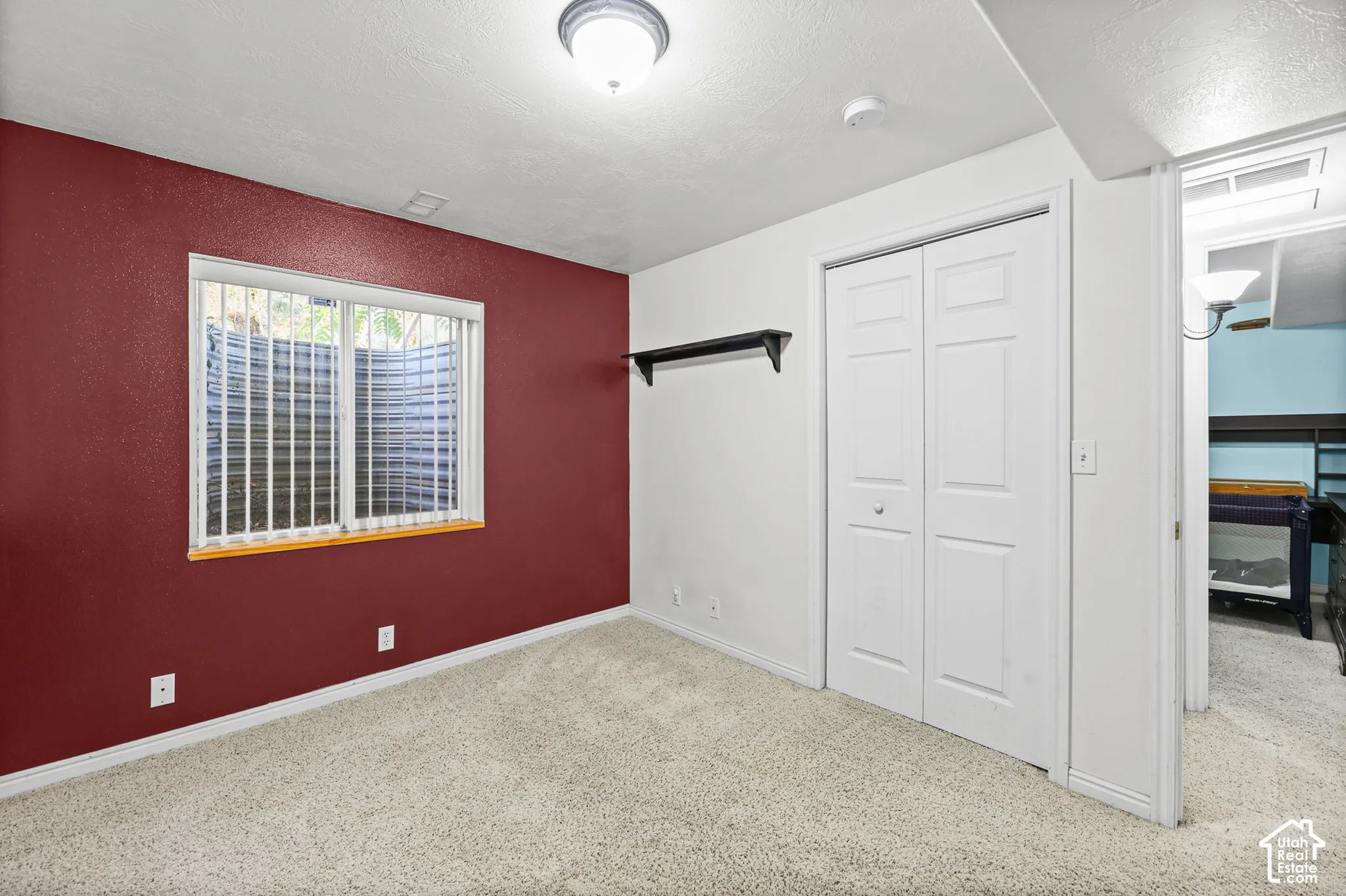 Window with vertical blinds, light-colored carpeting, white bi-fold closet doors, wall-mounted shelving, and ceiling-mounted light fixture