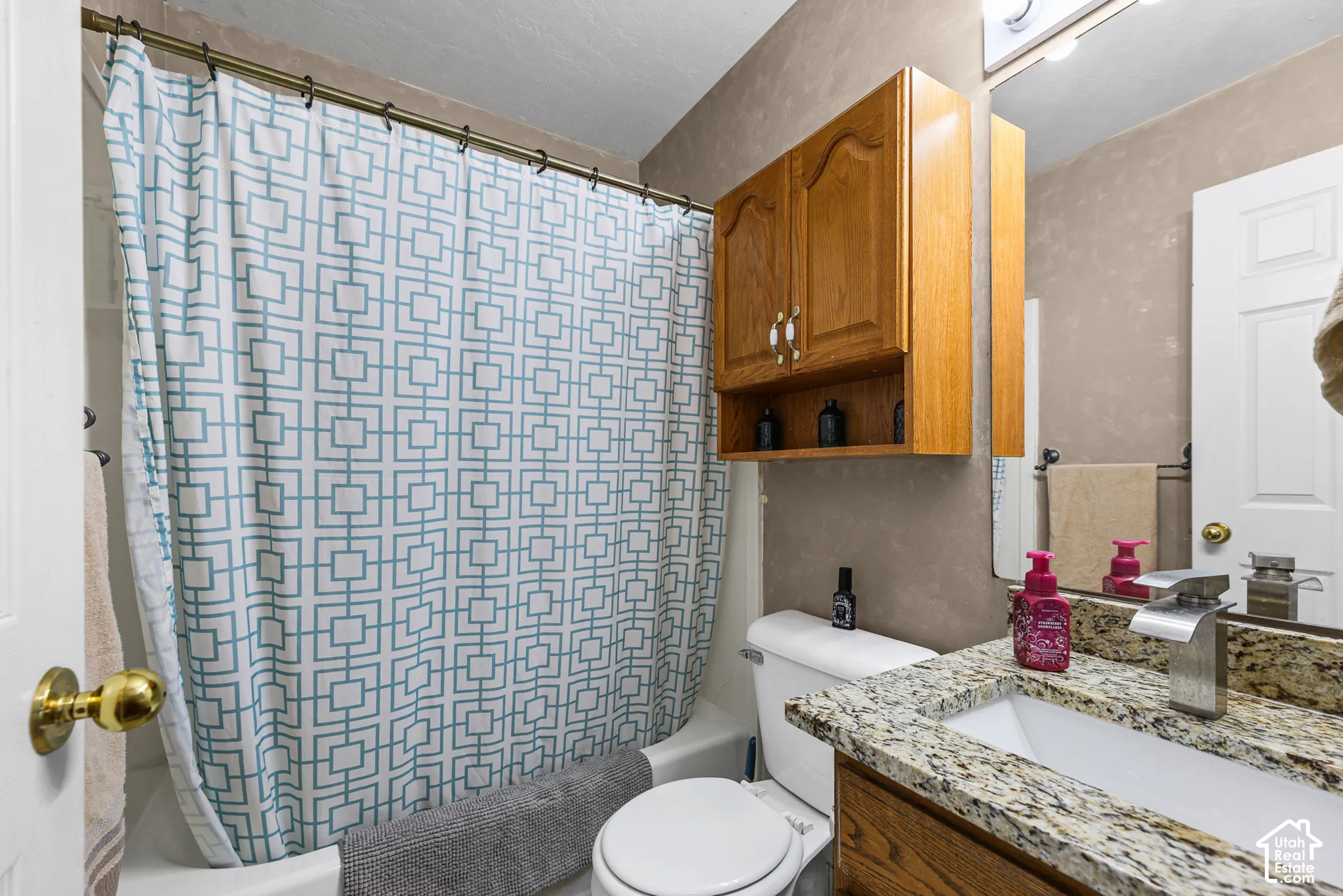 Bathroom featuring a built-in tub with shower, wood-finish vanity with a granite-style countertop, integrated sink, and an overhead wood-finish cabinet with shelving