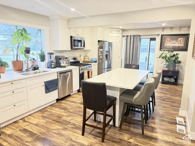 Kitchen featuring wood-finish flooring, white cabinetry, a large window over the sink, stainless steel appliances, and a central island with seating