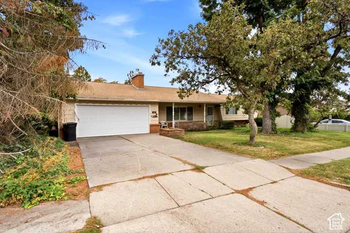 Single-story residence featuring a brick chimney, an attached garage, a concrete driveway, and established landscaping