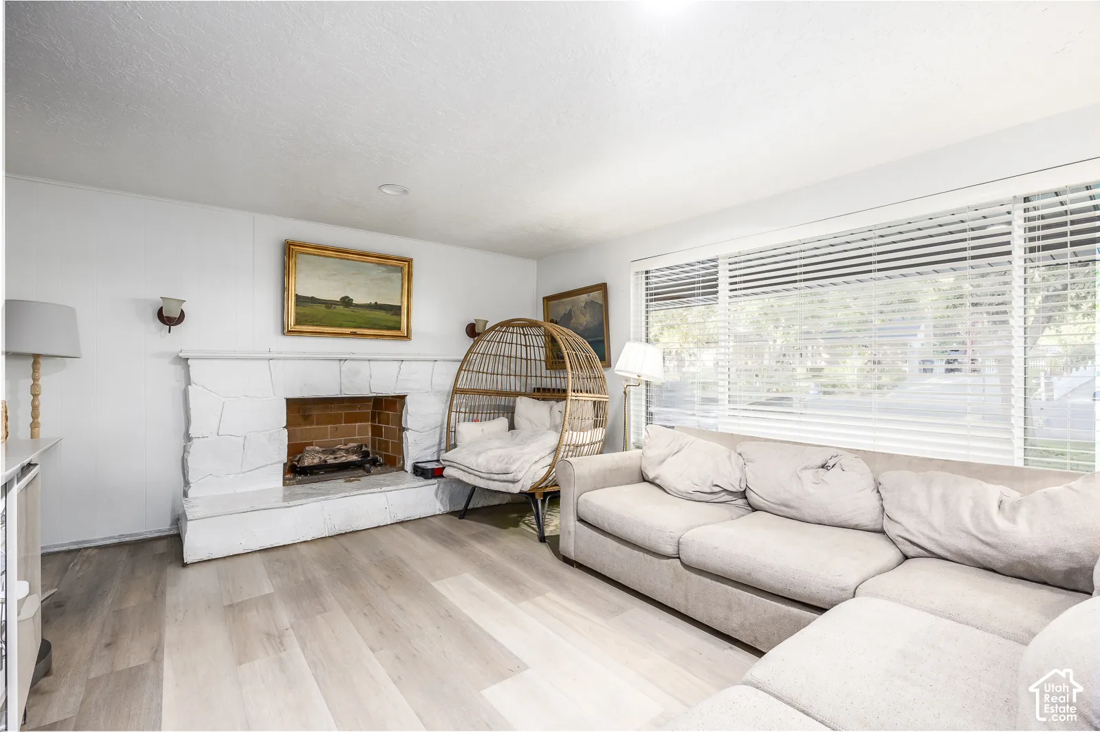 Light wood-finish flooring, stone-clad fireplace with brick firebox, wall-mounted sconces, and extensive window with horizontal blinds