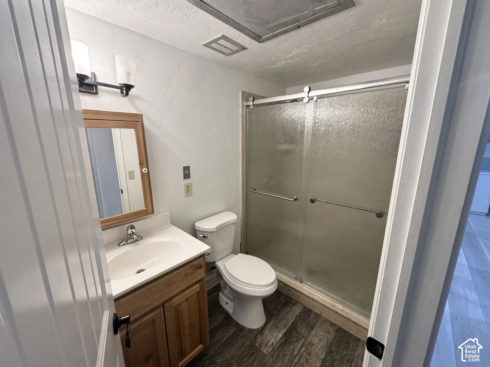 Wood-finish flooring, single vanity with wood cabinetry, glass-enclosed shower with sliding doors, wall-mounted light fixture, and a framed mirror