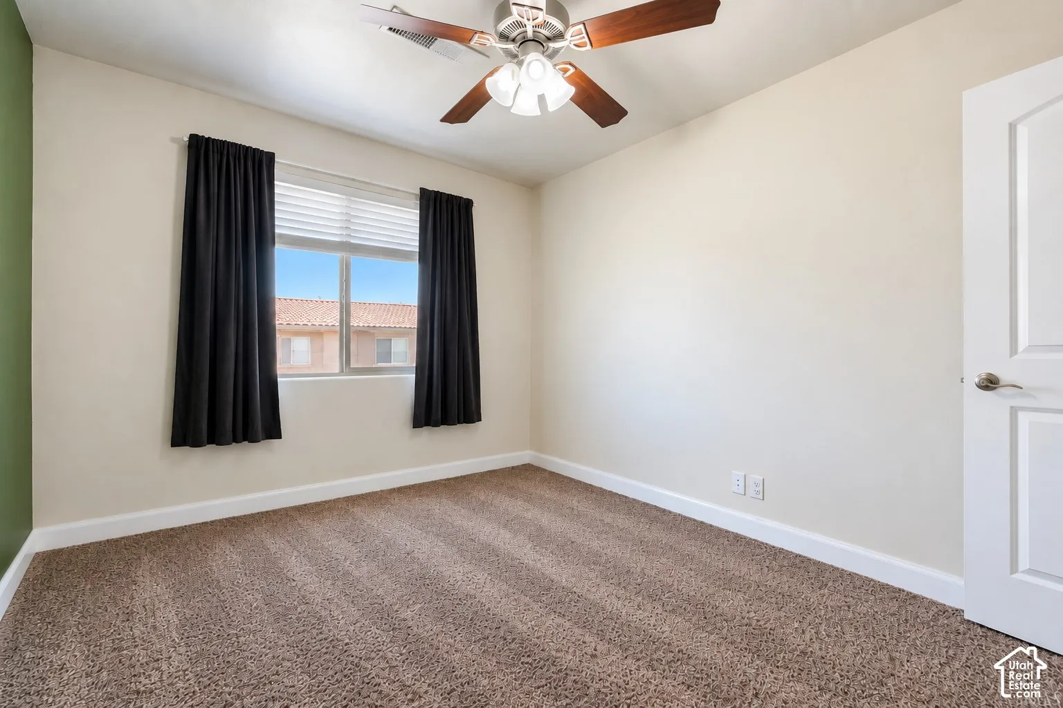Ceiling fan with wood-finish blades, window with blinds and dark curtains, neutral tone carpeting, white baseboards, and a white interior door with brushed nickel hardware
