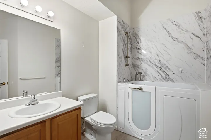 Bathroom featuring a vanity with wood-finish cabinetry and white countertop, walk-in tub with a shower fixture, and a marble-patterned wall surround