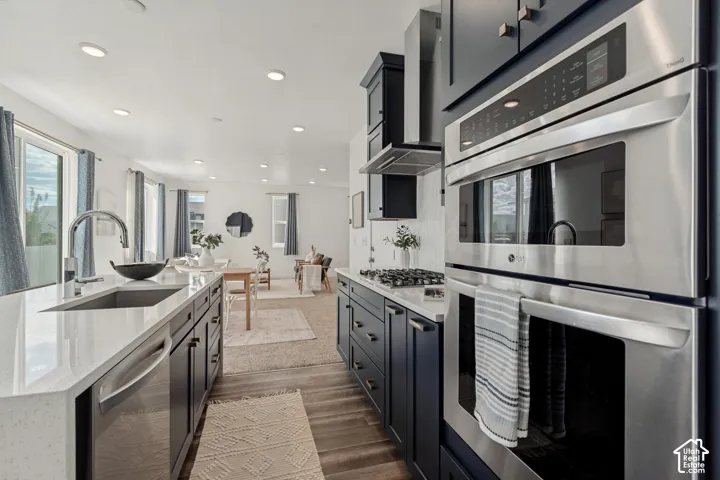 Kitchen featuring a large island with an undermount sink, stainless steel appliances including a built-in oven and microwave, dark cabinetry, and wood-finish flooring