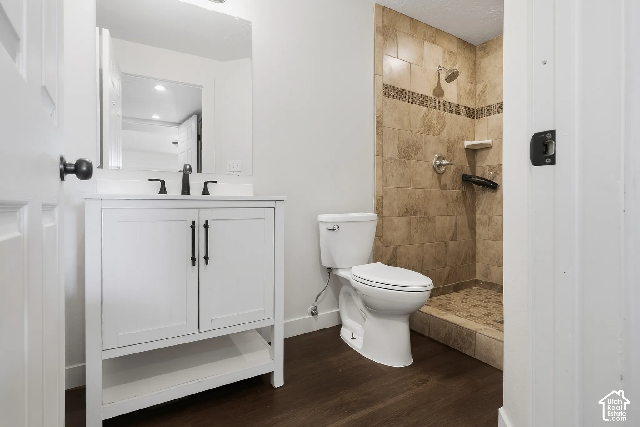 White vanity with black hardware and faucet, wood-finish flooring, and a tiled shower with an accent band