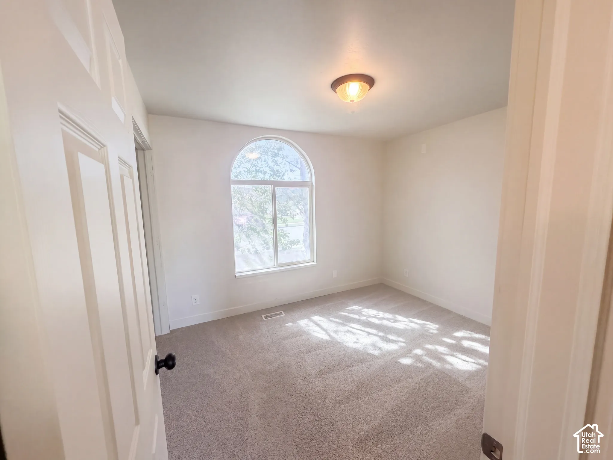 Arched window with clear glass, light-colored carpet flooring, flush-mount ceiling light fixture, white interior walls, and white paneled door