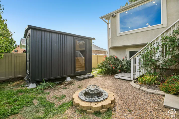 Detached auxiliary structure with vertical siding and glass door, gravel hardscaping, permanent stone fire pit, wood staircase with white railing, and stucco exterior with large window