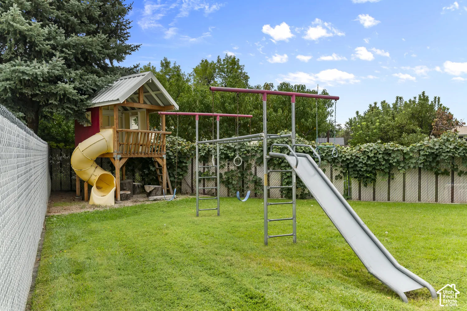 Expansive backyard with green lawn, chain link fencing, mature trees, and a built-in wooden play structure with a slide
