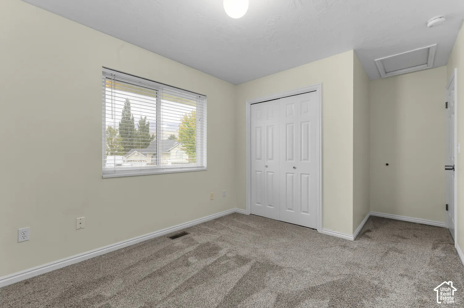 Neutral wall color, carpeted flooring, white bi-fold closet doors, window with horizontal blinds, and a flush-mount ceiling light