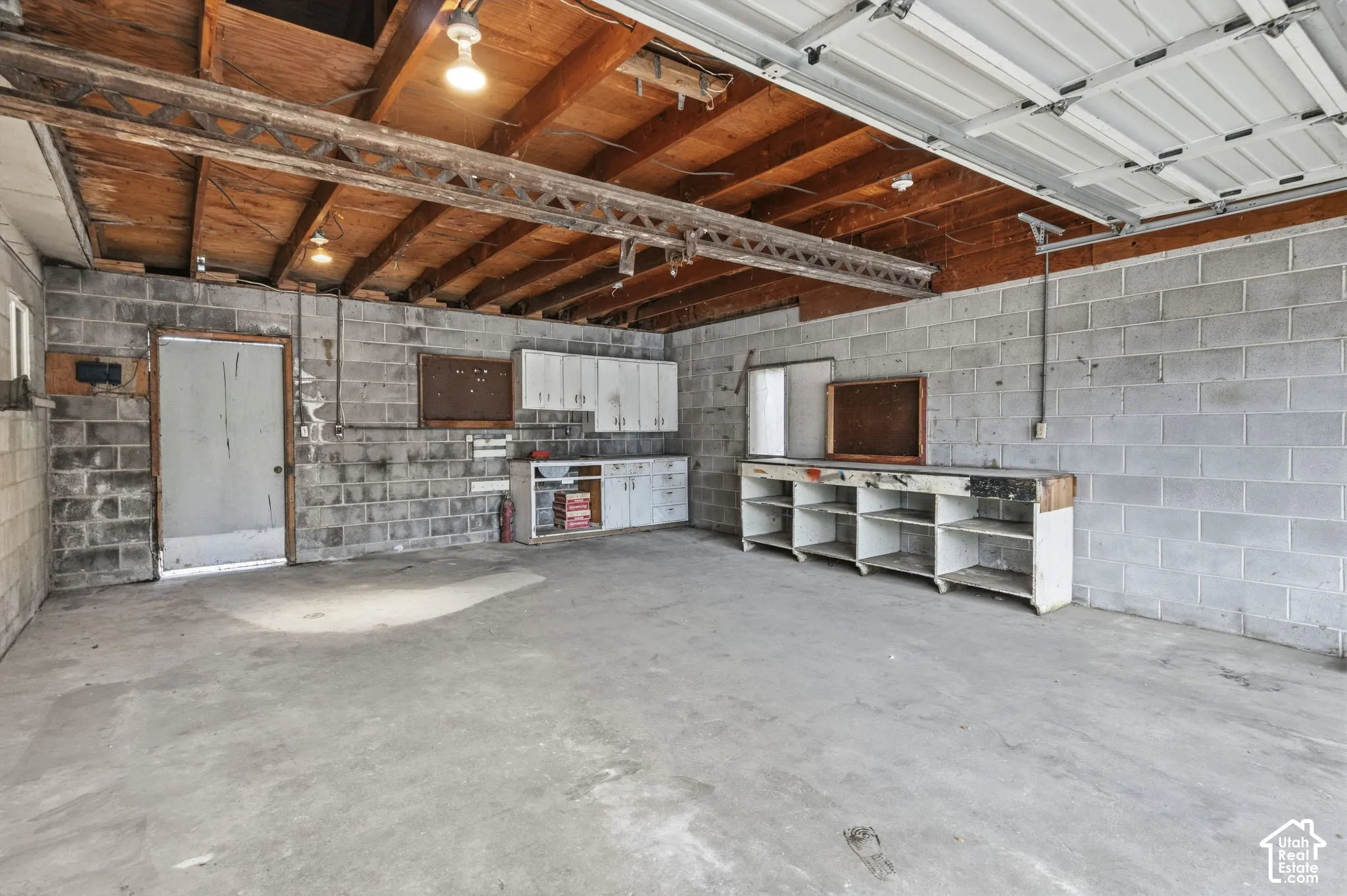 Garage attached to Detached Building - Concrete block walls with exposed wood ceiling beams, concrete floor, built-in shelving, and overhead lighting, bridge beam for engine hoist