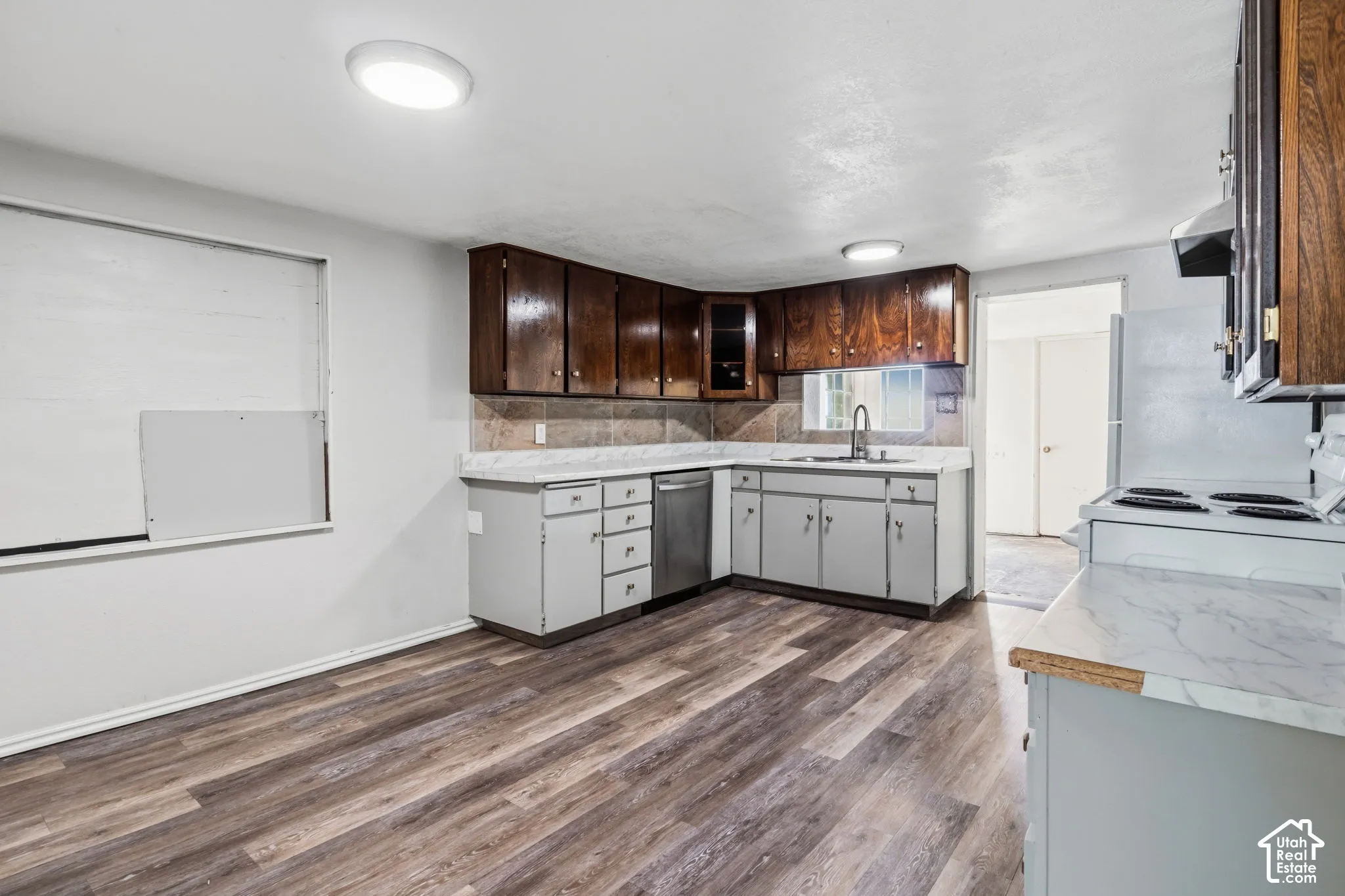 Laminate flooring, dark wood upper cabinetry, light gray lower cabinetry, light-toned countertops, and a stainless steel dishwasher. large in-kitchen dining space, window space covered.