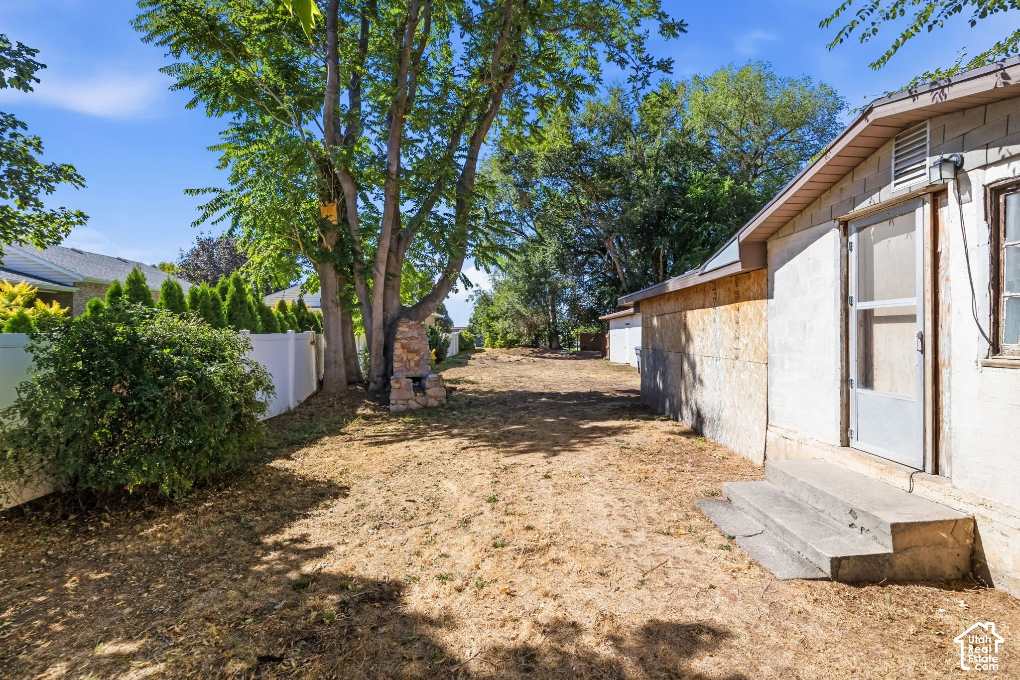 Rear exterior featuring mature trees, a stone-clad fireplace, and a white privacy fence, back door to laundry