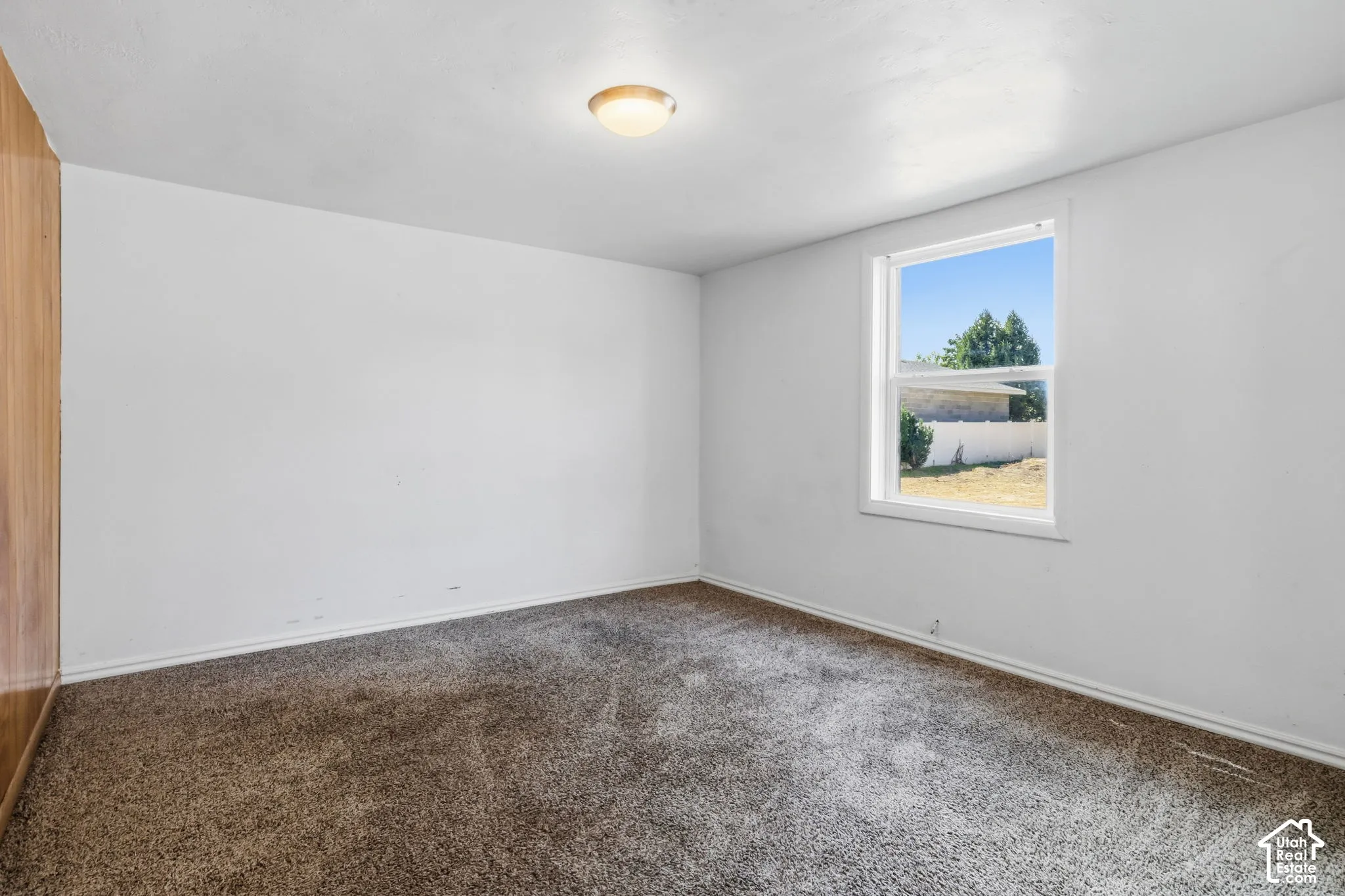 White interior walls, single window, overhead lighting, brown/gray carpet flooring, wood-paneled wall