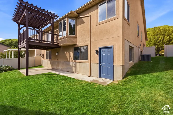 Raised wood deck with pergola, lower concrete patio, stucco exterior, multiple windows, and a blue exterior door