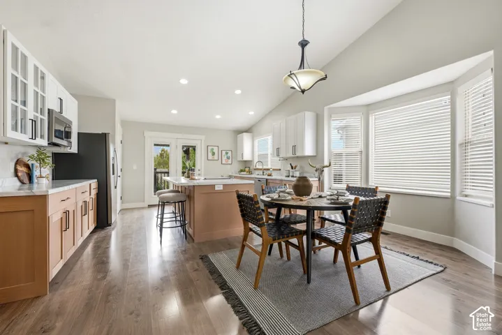 Wood-finish flooring, vaulted ceiling, white upper cabinetry with glass inserts, light wood-finish lower cabinetry, and a bay window