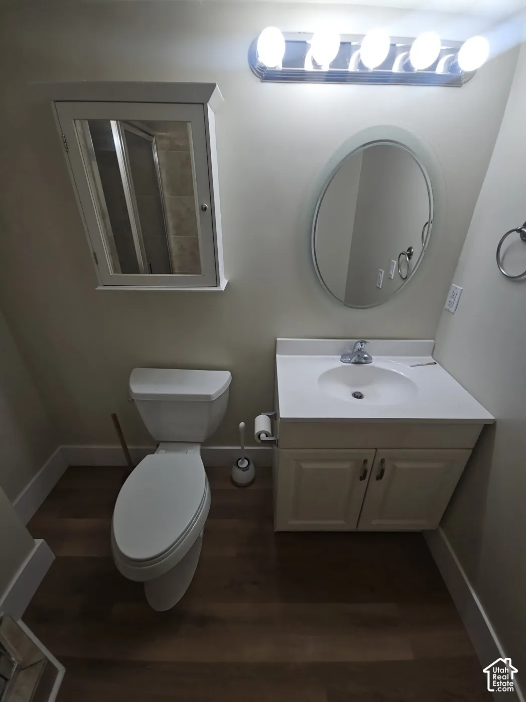 Bathroom featuring a vanity cabinet with a white countertop and integrated sink, oval wall-mounted mirror, recessed medicine cabinet, and wood-finish flooring