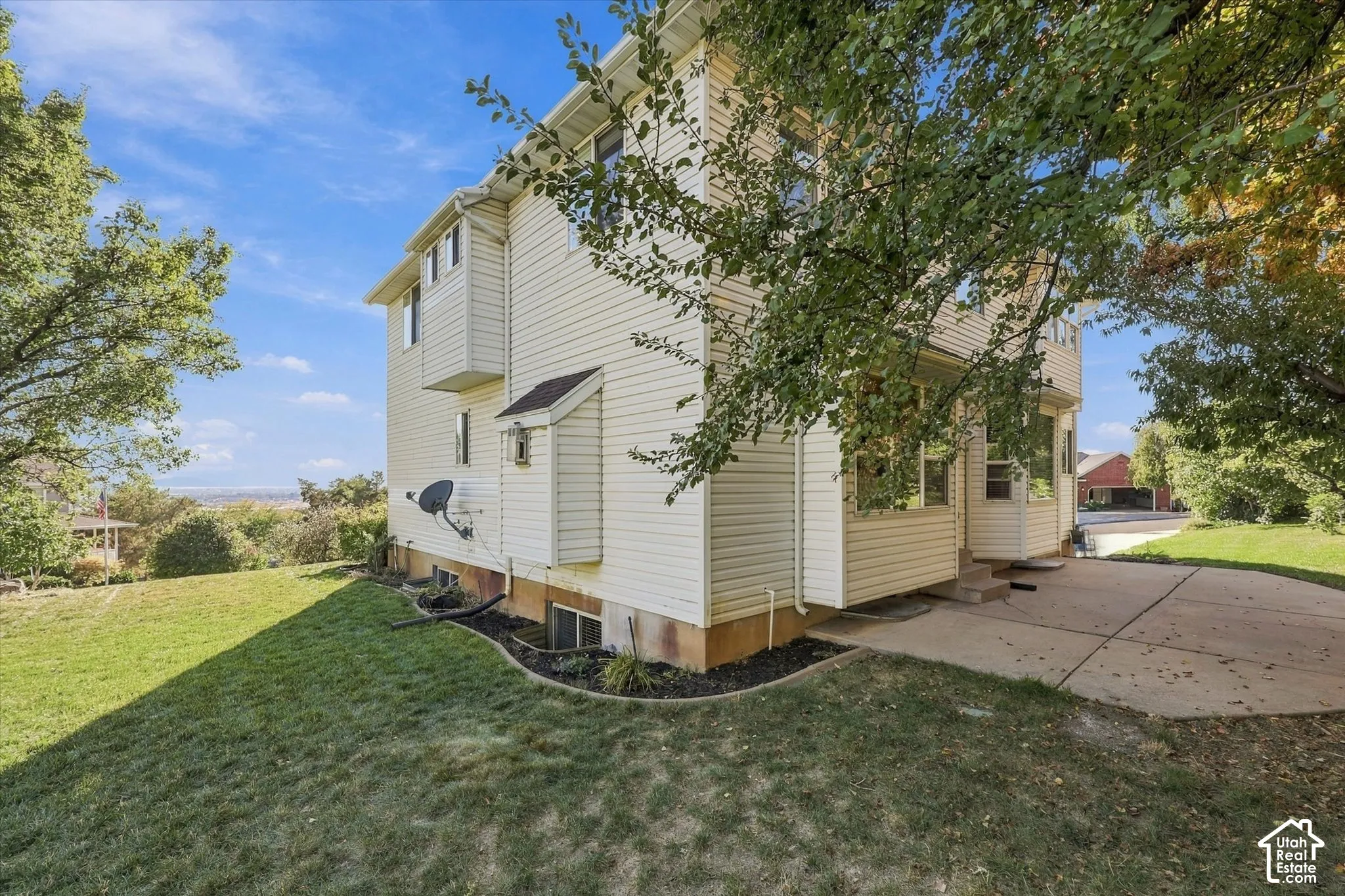 Side and Back yards. Cream-colored siding exterior with multiple windows, concrete patio, sloped grassy area, and mature trees