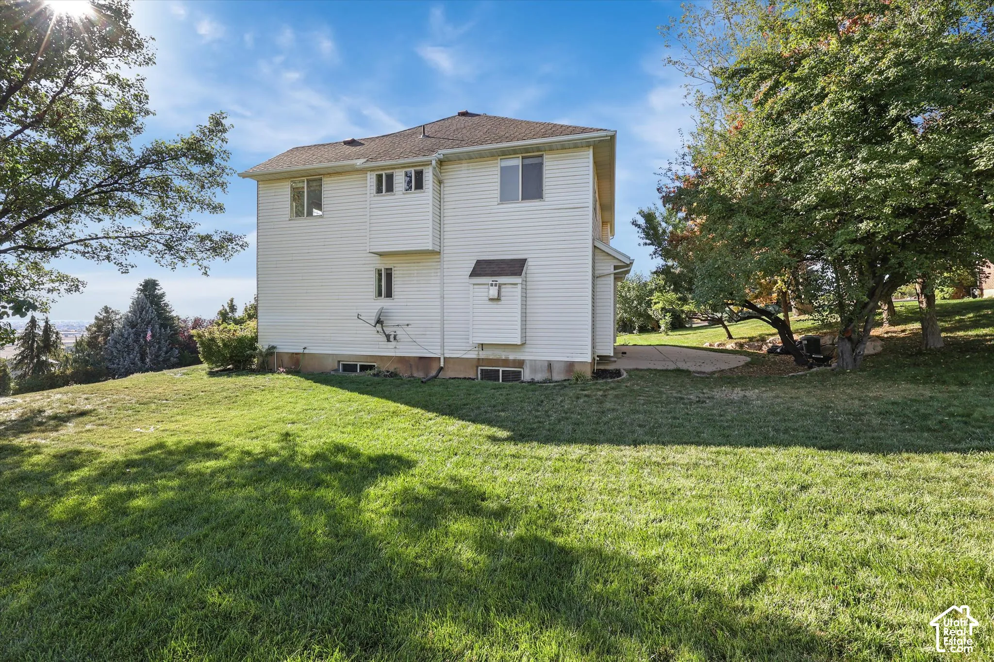 Large side yard on cul-de-sac. White multi-level property with a brown shingle roof, surrounded by mature trees and a sloped lawn