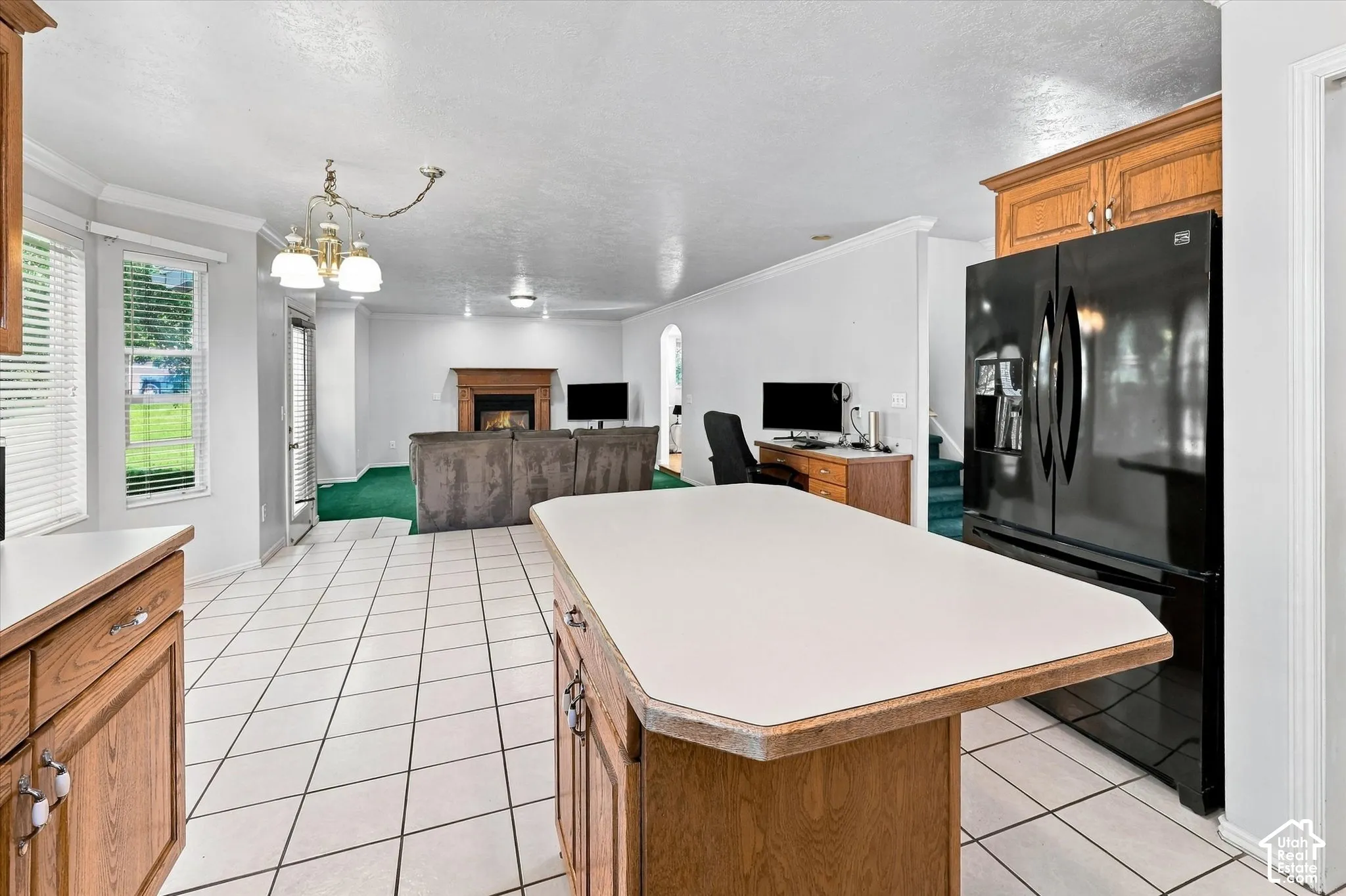 White tile flooring, wood-finish cabinetry, central island with solid surface countertop, black refrigerator, and a fireplace with a wood mantel