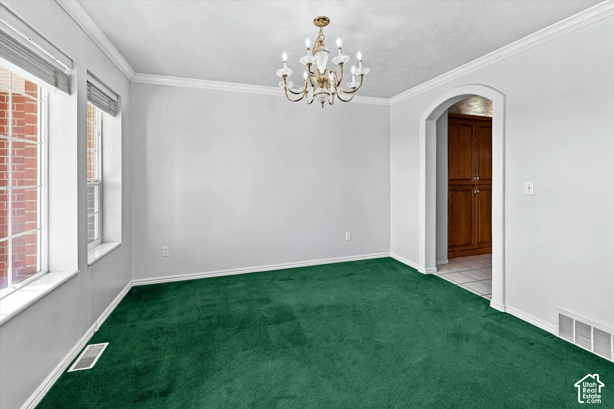 Elegant formal dining room featuring pristine white walls, crown molding, a brass chandelier with candle-style arms, large windows with natural light, arched doorway with wood-finish door, and rich emerald carpet flooring