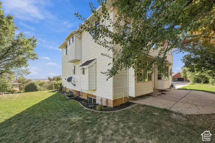 Side and Back yards. Cream-colored siding exterior with multiple windows, concrete patio, sloped grassy area, and mature trees