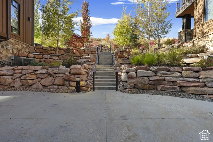 Concrete pathway leading to a stone staircase with metal handrails, flanked by terraced stone retaining walls and mature landscaping