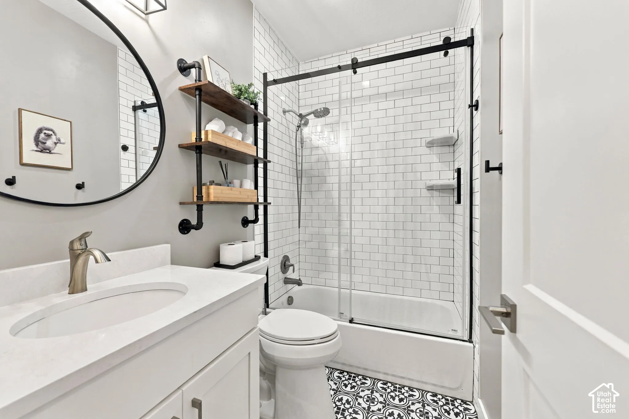 White vanity with a single basin sink and brushed nickel faucet, round framed mirror, black and white patterned floor tile, subway tile shower surround, and sliding glass shower door with black hardware