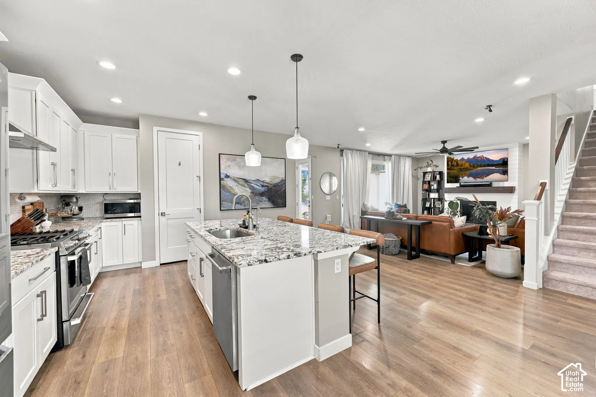 Open-concept living space with wood-finish flooring, a kitchen island with seating, white cabinetry, stainless steel appliances, and a staircase with a wood handrail