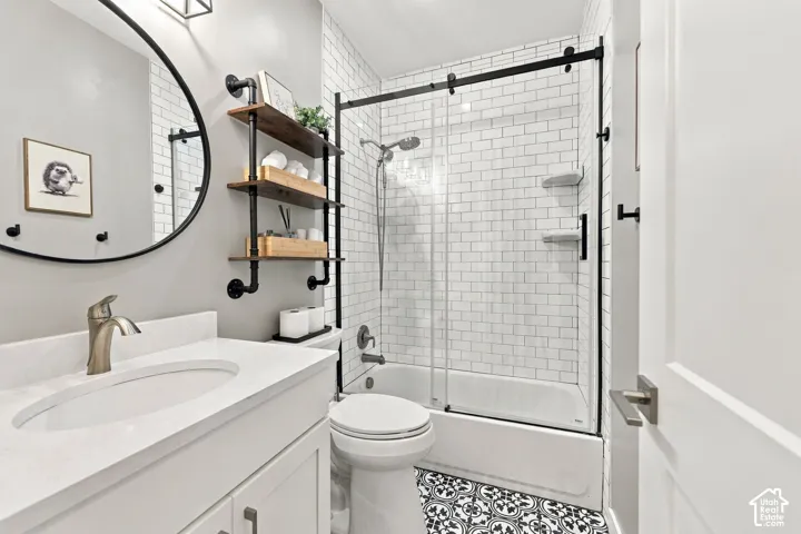 White vanity with a single basin sink and brushed nickel faucet, round framed mirror, black and white patterned floor tile, subway tile shower surround, and sliding glass shower door with black hardware
