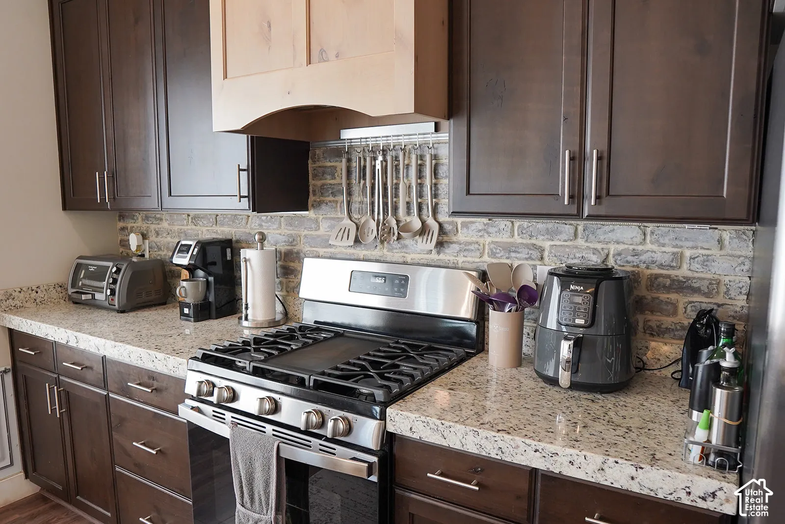 Dark wood-finish cabinetry with silver-tone hardware, light-toned countertop, stainless steel range with griddle, wood-finish range hood, and brick-patterned backsplash