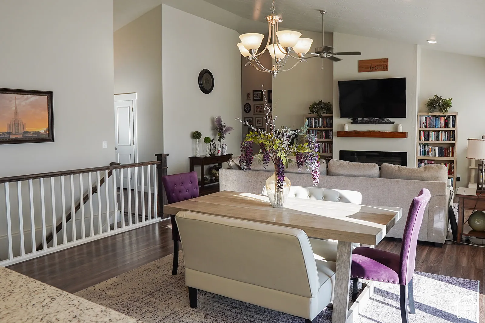 Vaulted ceiling with an integrated ceiling fan and chandelier, dark wood-finish flooring, white spindle stair railing, and a fireplace with an integrated mantel