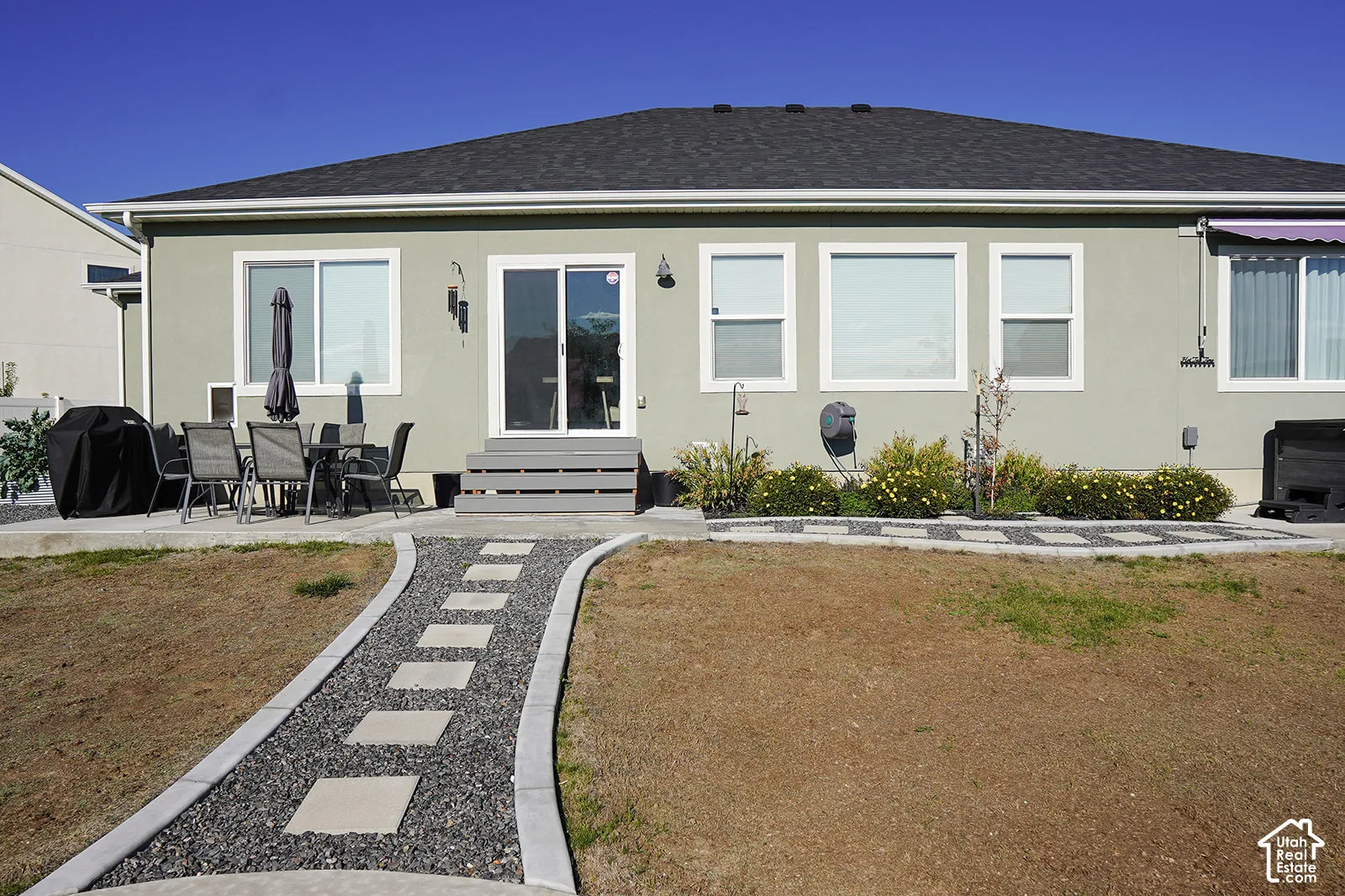 Rear exterior with a concrete patio, sliding glass door, multiple windows, and a paved walkway featuring square stepping stones and decorative gravel