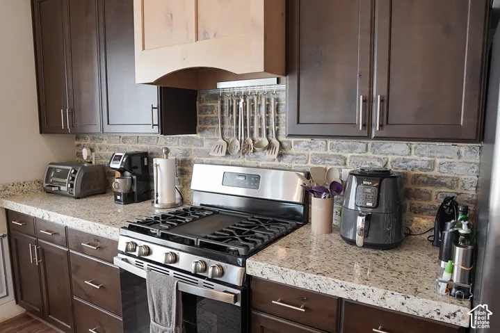 Dark wood-finish cabinetry with silver-tone hardware, light-toned countertop, stainless steel range with griddle, wood-finish range hood, and brick-patterned backsplash