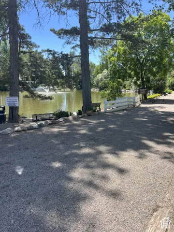 Gravel pathway leading to a tranquil pond with a decorative fountain, bordered by mature trees and a white picket fence