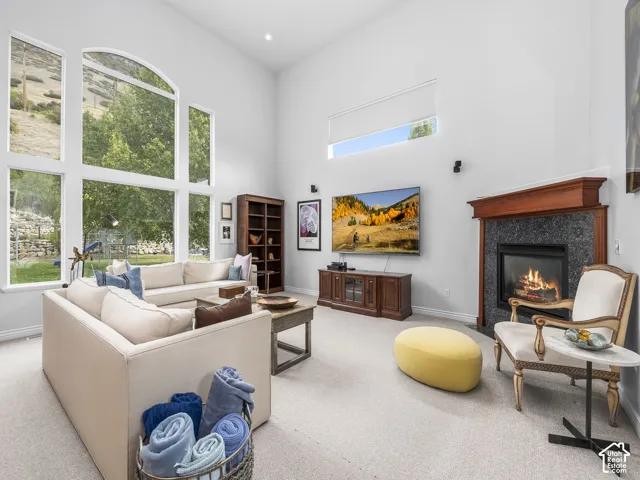 Spacious living area featuring high ceilings, a grand fireplace with a dark stone surround and wood mantel, expansive multi-pane windows, and light-colored carpeting