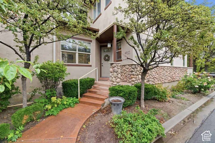 Stucco exterior with river rock accents, covered entryway, mature trees, shrubbery, and a concrete pathway leading to the entrance