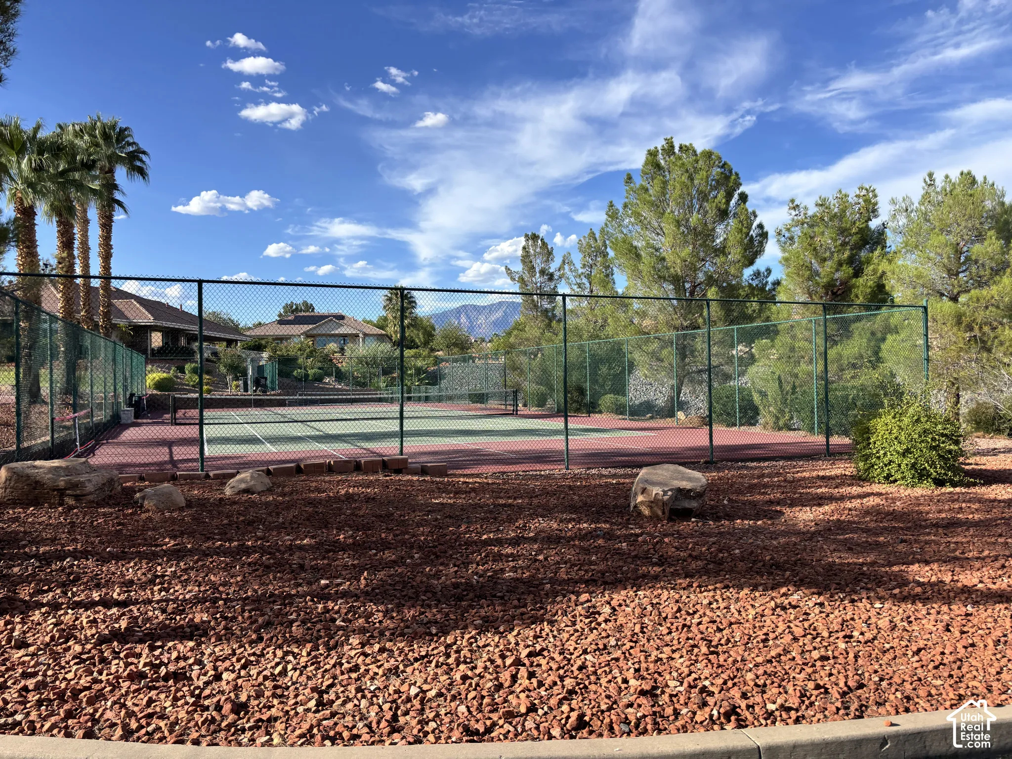 Tennis court with perimeter fencing, surrounded by xeriscaped landscaping featuring red rock and palm trees