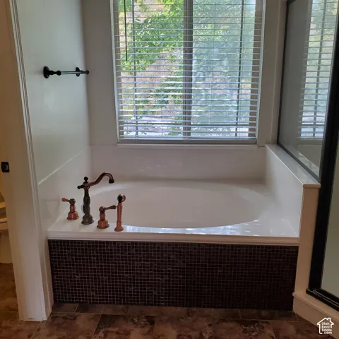 Integrated soaking tub with mosaic tile surround, copper-toned fixtures, and window with privacy blinds