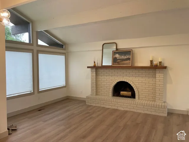 Vaulted ceiling with exposed beams, light wood-finish flooring, white brick fireplace with an arched firebox, wood mantel, and multiple large windows
