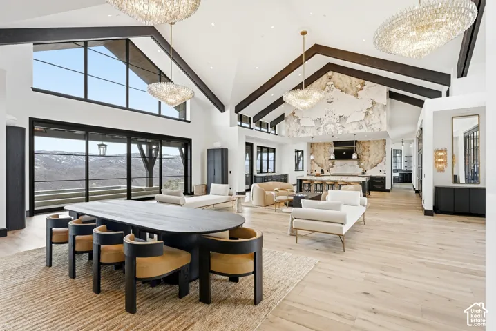 Vaulted ceilings with exposed beams, light wood-finish flooring, expansive window walls, two chandelier light fixtures, and a statement wall with textured stone finish