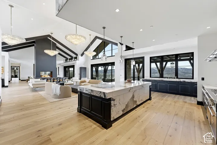Expansive open-concept space featuring wood-finish flooring, vaulted ceilings with exposed beams, a large stone-clad fireplace, a substantial kitchen island with a stone countertop, and black-framed windows with mountain scenery