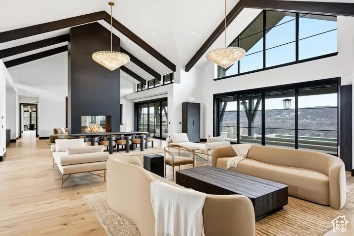 Vaulted ceilings with exposed beams, wood-finish flooring, a contemporary fireplace with a stone hearth, floor-to-ceiling windows, and elegant chandeliers